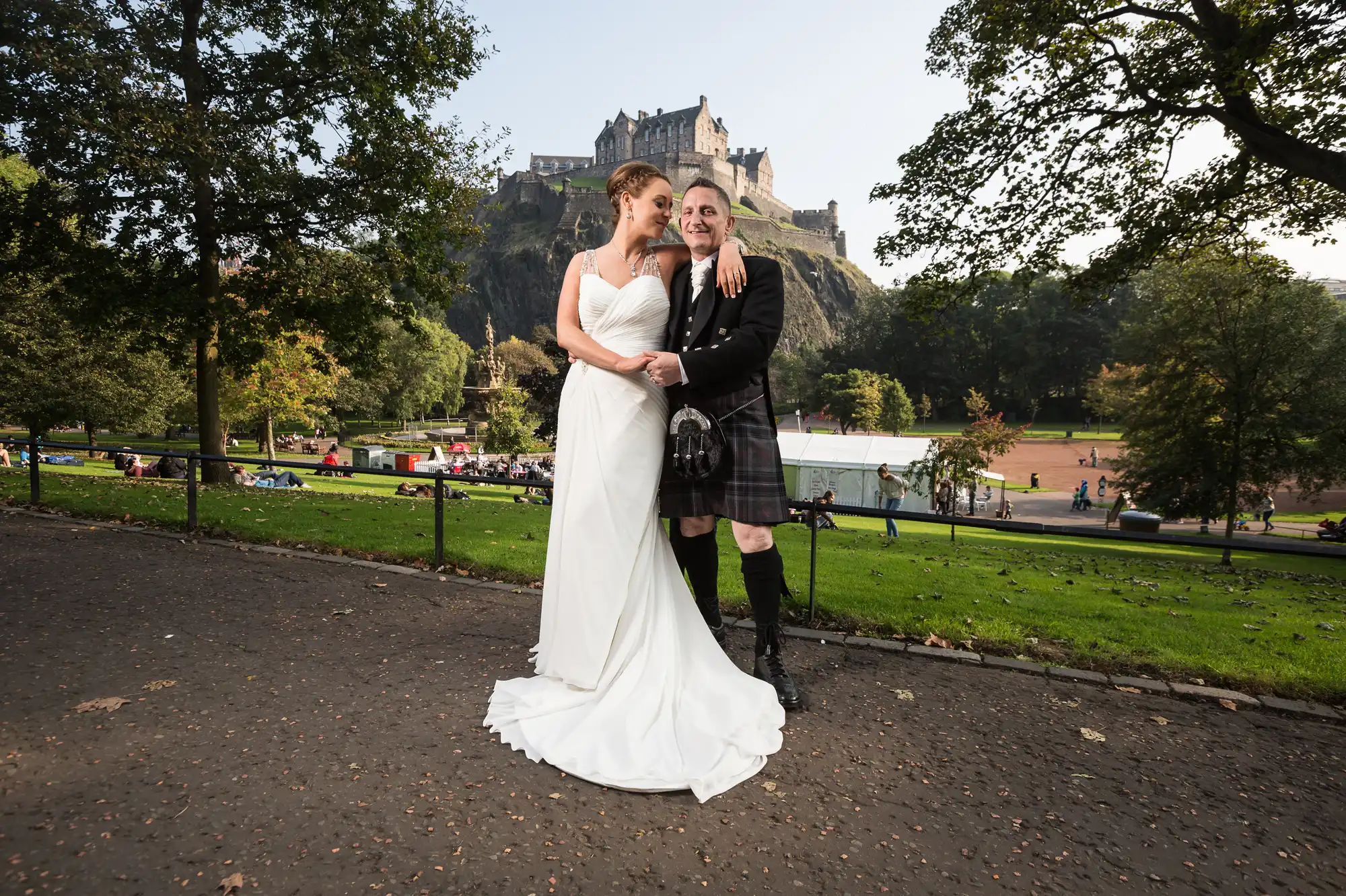 A bride in a white dress and a groom in a kilt embrace in a park with edinburgh castle in the background.