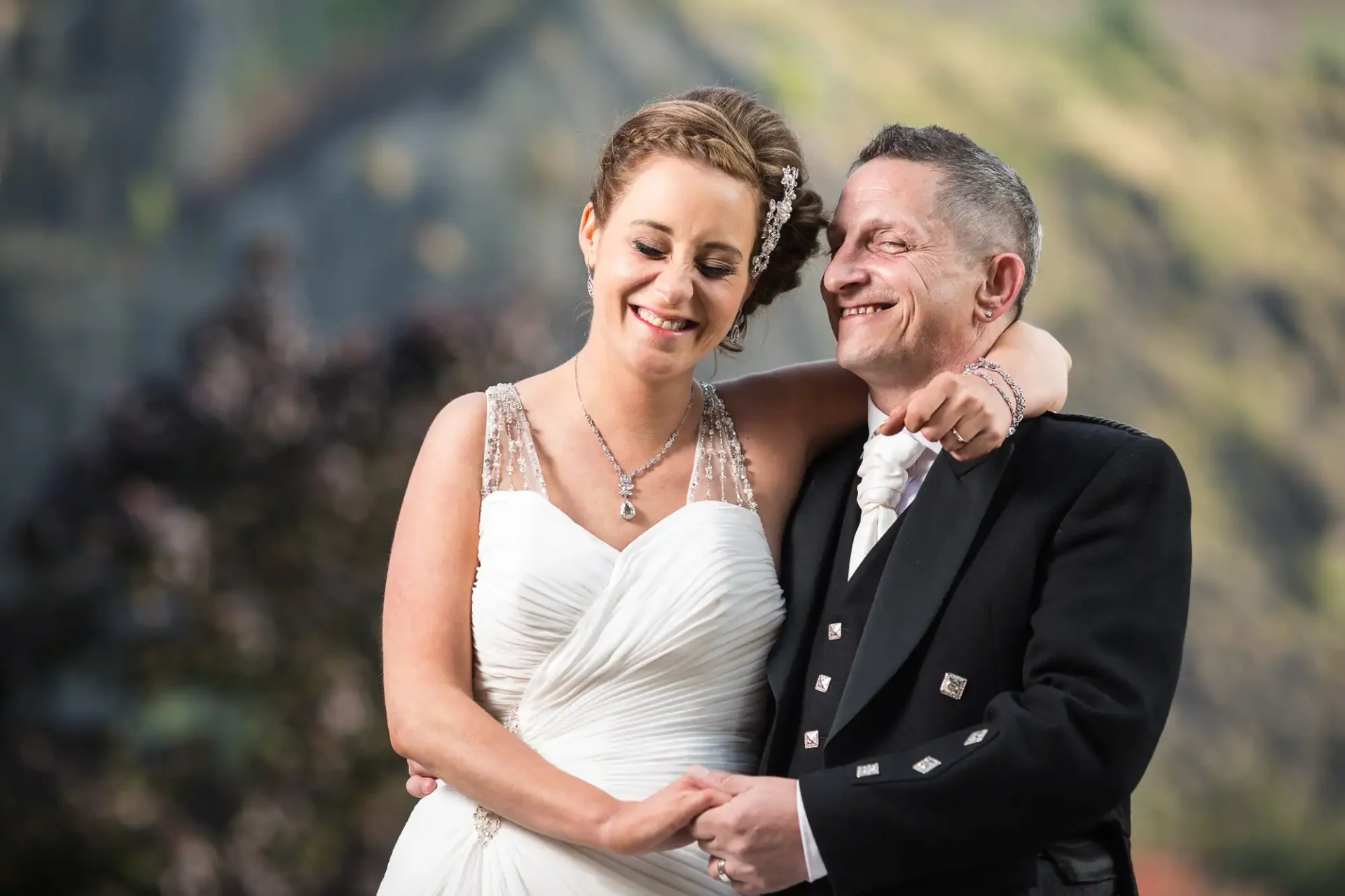 Ghillie Dhu wedding photos: A bride and groom smiling and embracing outdoors, with a scenic mountain backdrop. the bride wears a white gown and the groom is in a black tuxedo.