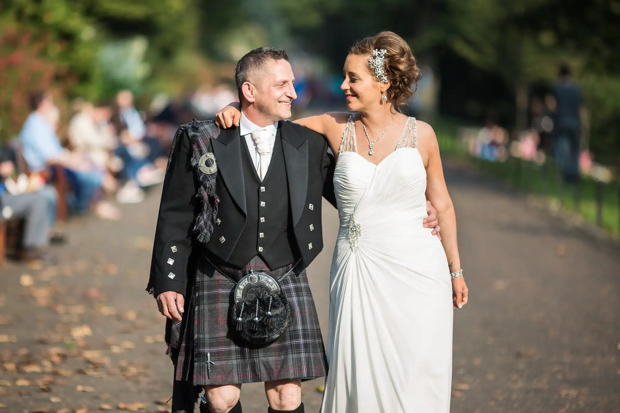 A newlywed couple joyfully walking in a park, with the man in a traditional kilt and the woman in a white wedding gown.