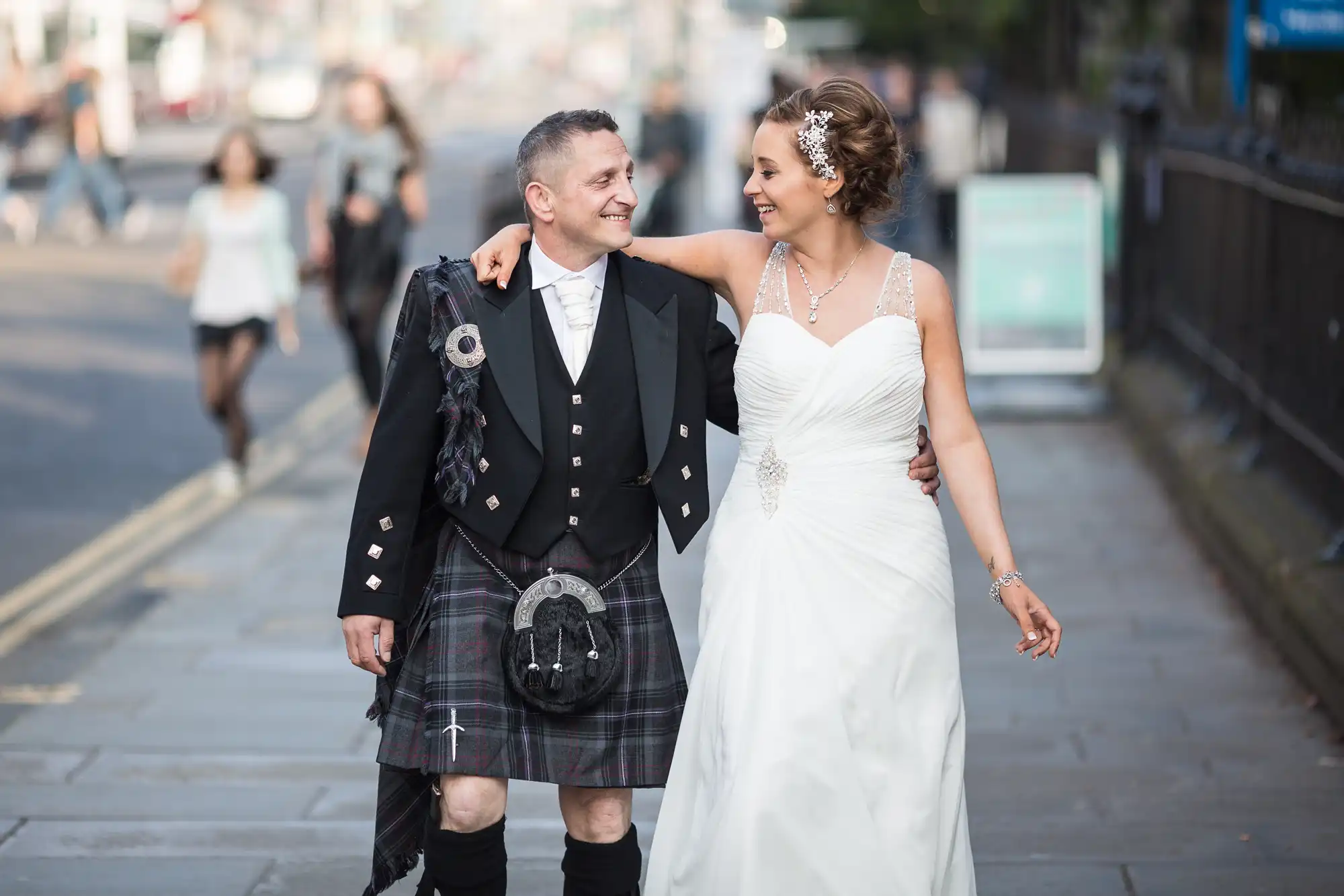 Bride in a white dress and groom in a kilt walking on a city street, smiling and looking at each other.