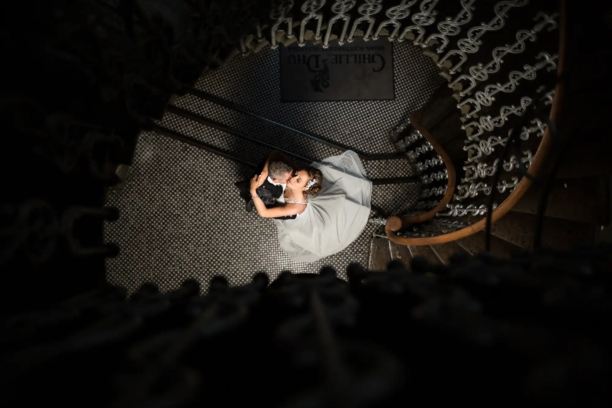 Wedding couple embracing on a spiral staircase from an overhead perspective, with intricate railing patterns surrounding them.