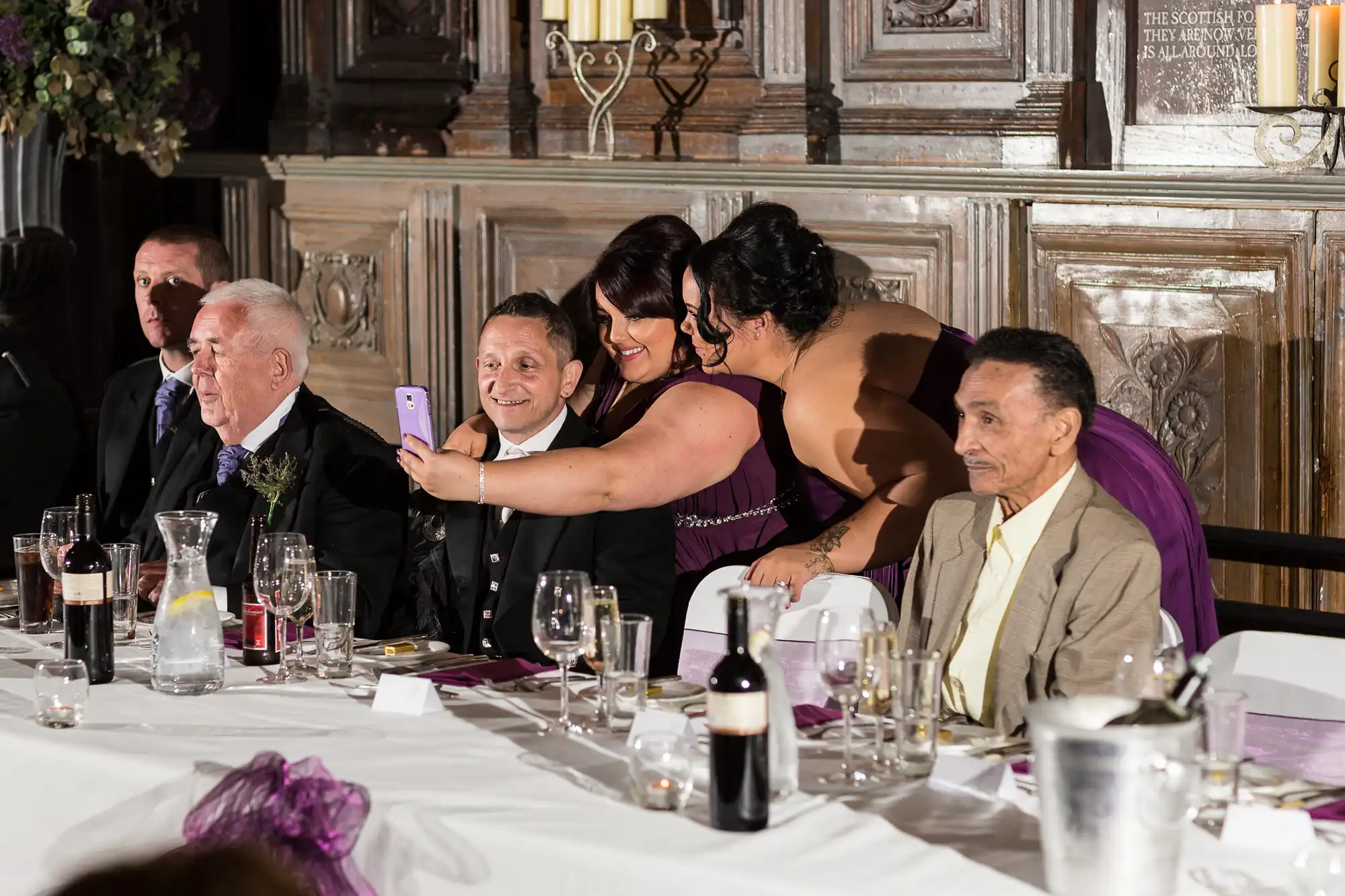 A group at a wedding banquet table takes a selfie, smiling, in an ornately decorated room with chandeliers and candles.