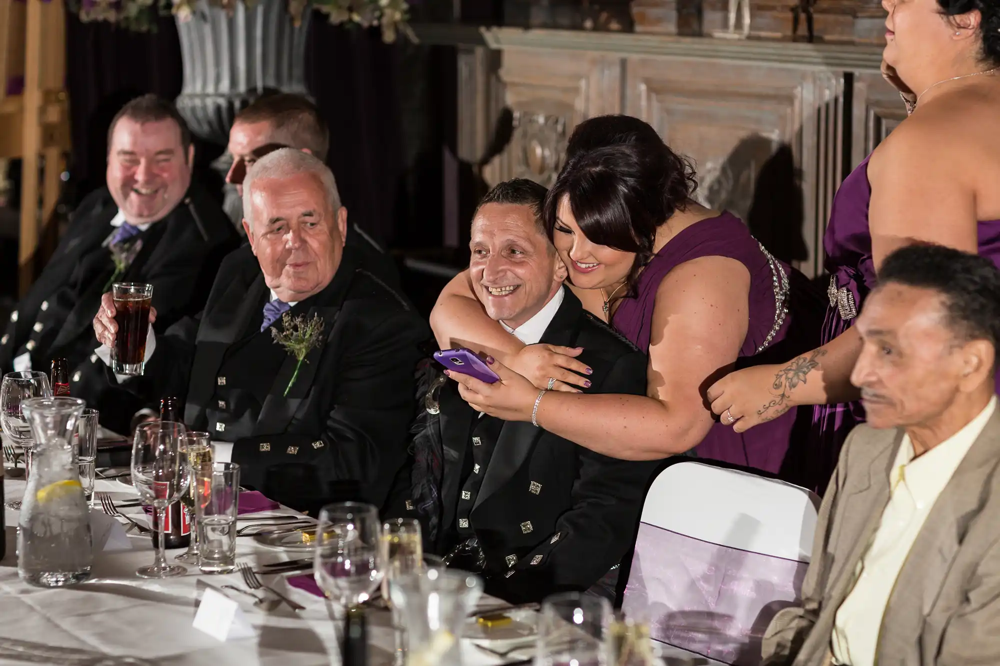 A group of guests at a formal event, one smiling woman showing something on a smartphone to a man next to her, other guests smiling and engaged in conversation.