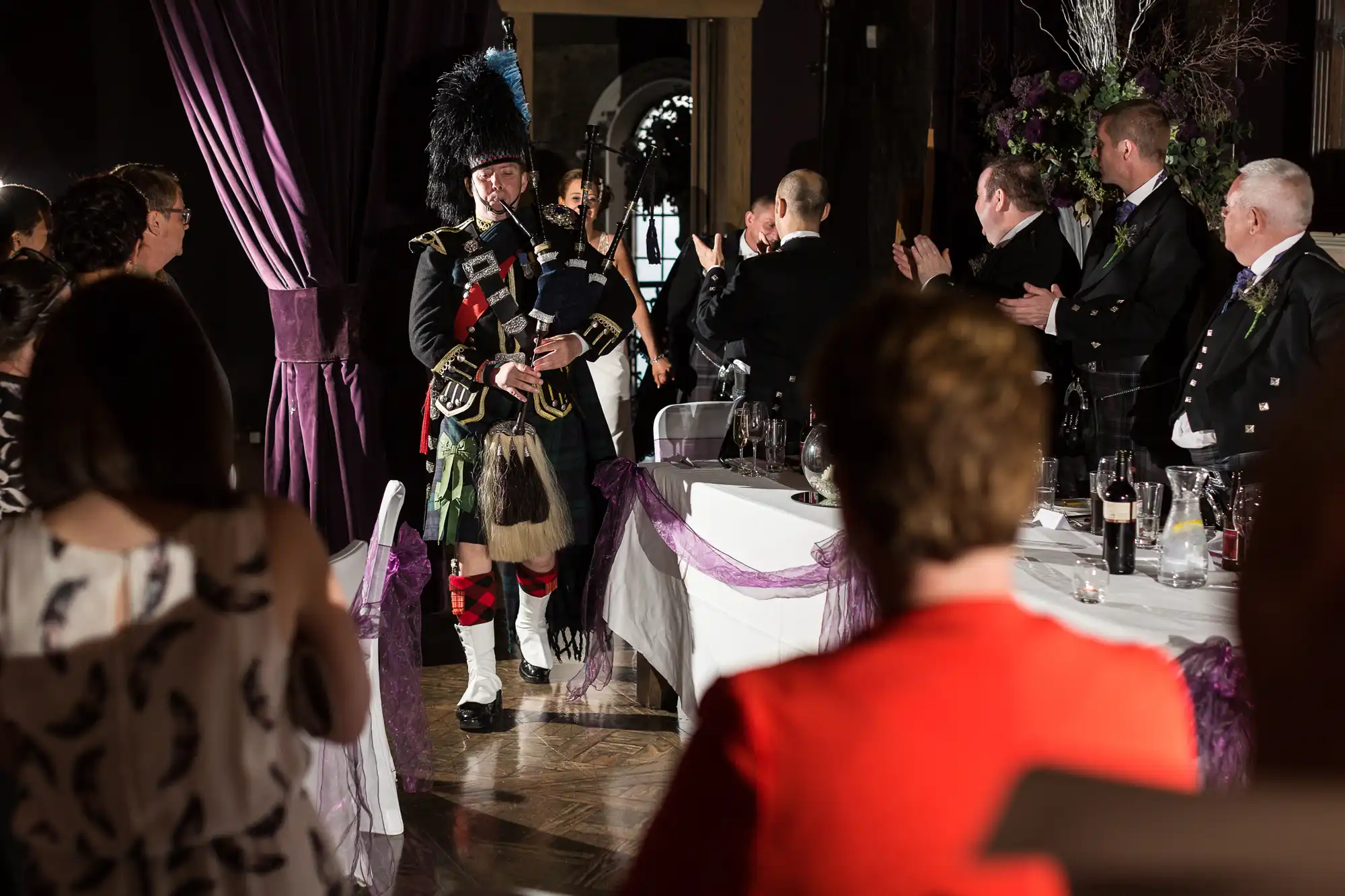 A bagpiper in traditional scottish attire performs at a wedding reception, surrounded by applauding guests.