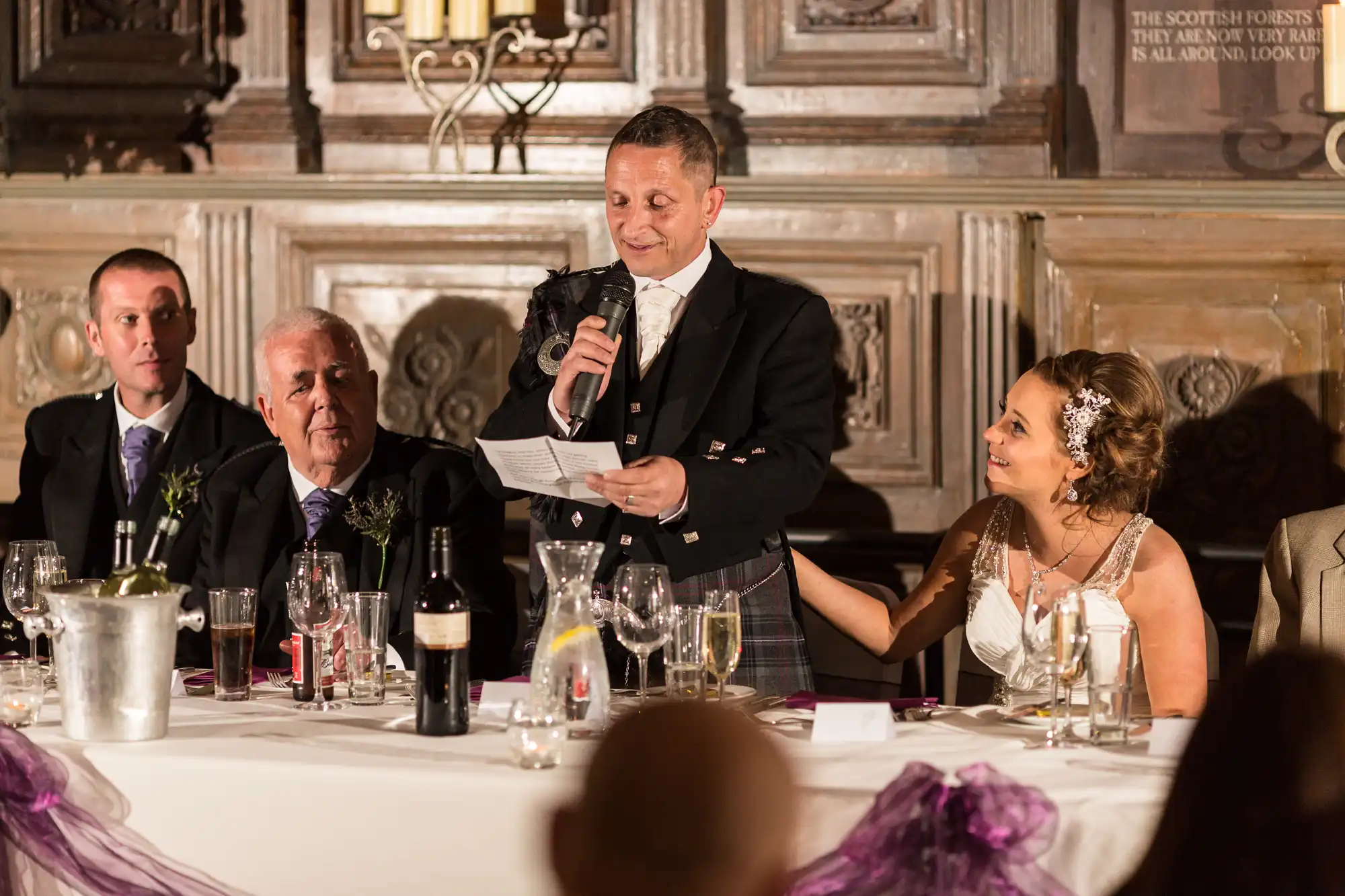 A man in a kilt gives a speech at a wedding reception while holding a microphone, with seated guests listening attentively.