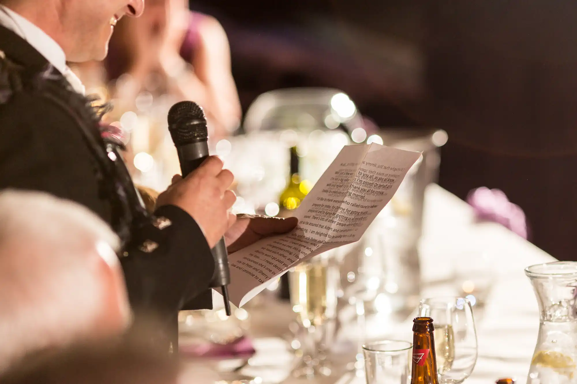A man in a suit giving a speech at a wedding reception, holding a microphone and a sheet of paper, with blurred guests in the background.