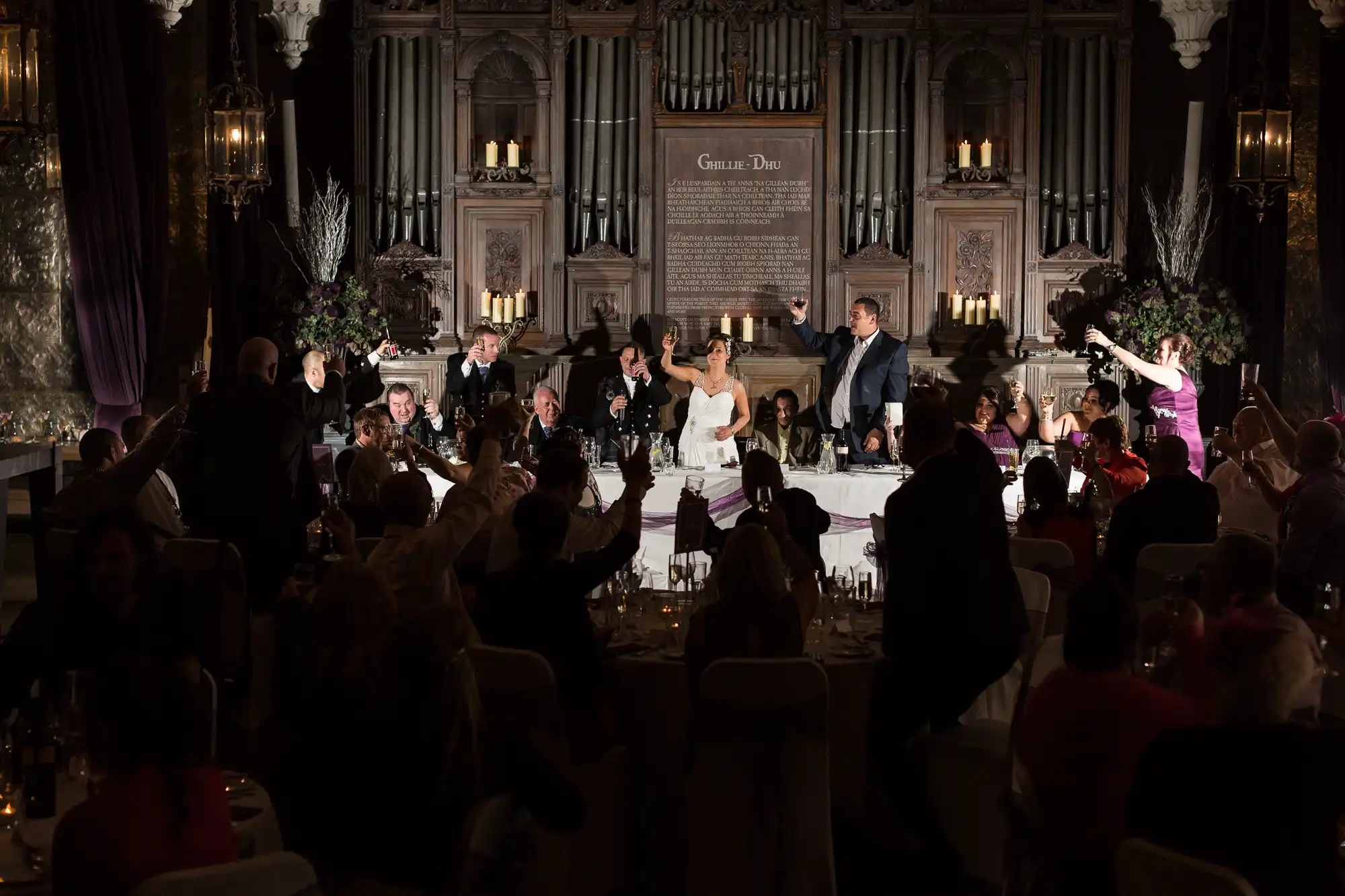 A bride and groom dancing on a table at an elegant, dimly lit wedding reception surrounded by cheering guests.