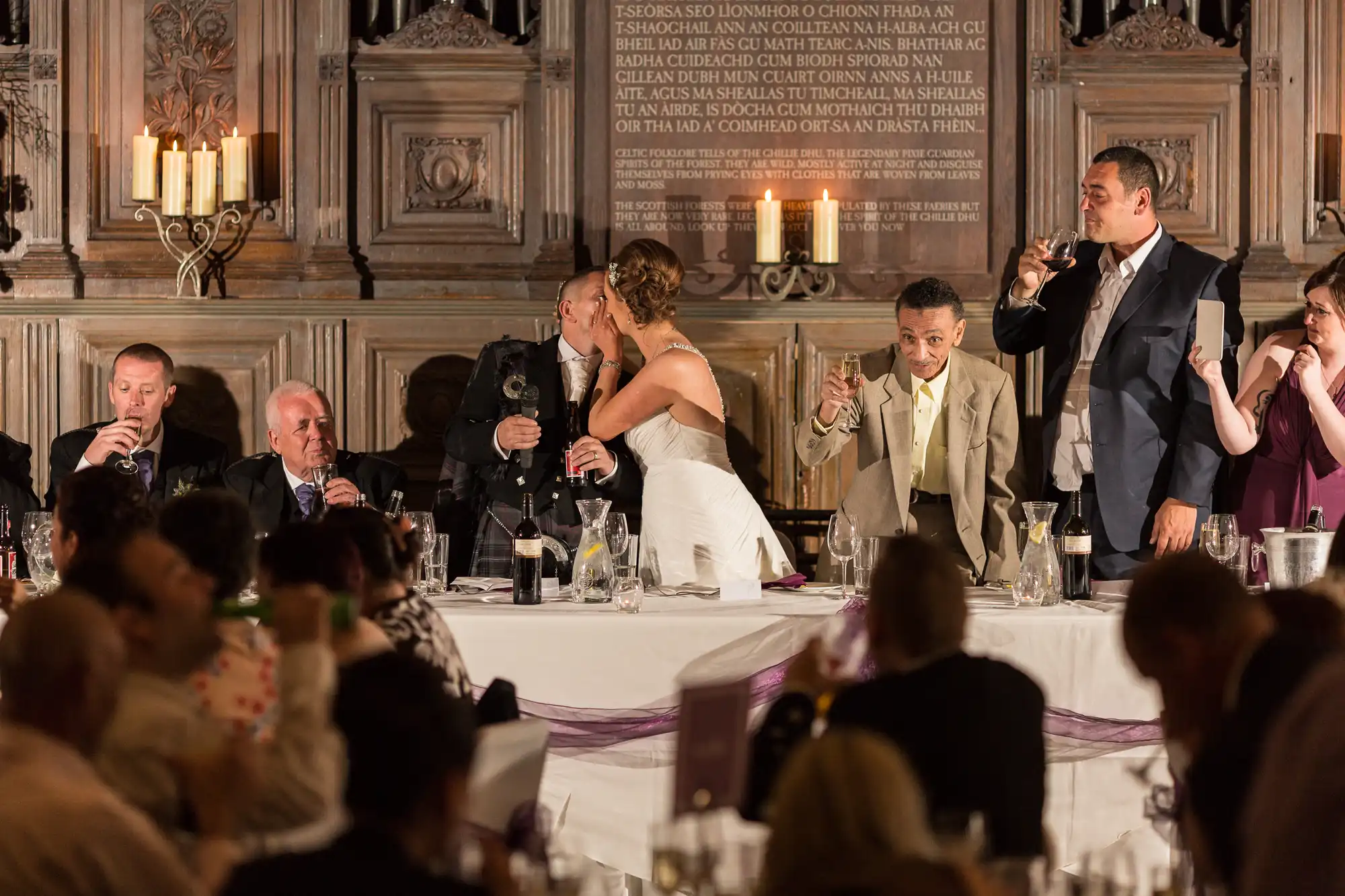 A bride and groom perform a toast at their wedding reception, surrounded by guests and ornate decor in a lavishly decorated room.