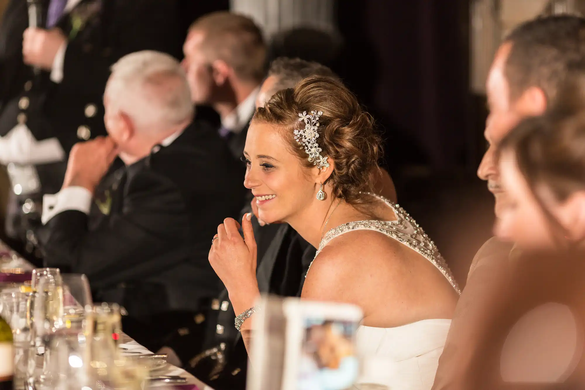 A bride in a beaded dress and floral hair accessory smiling during a banquet, with guests seated around her, blurred in the background.