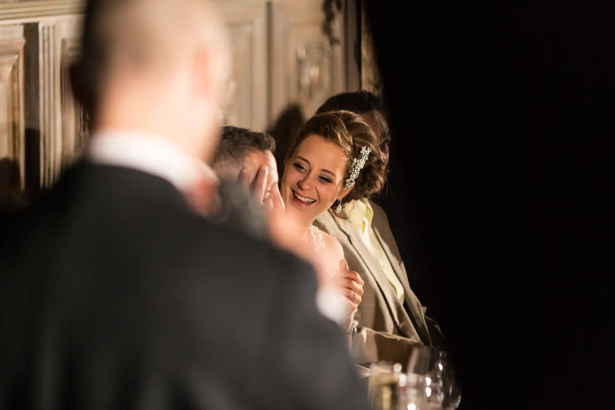 A bride joyfully looks at a groom during a wedding reception, both partially visible and warmly lit, with a focus on the bride's smiling face.