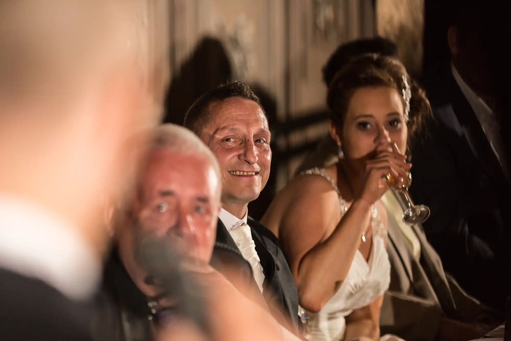 Man in a tuxedo smiling at a wedding reception, with other guests in foreground and beside him. focus is on the man in the center.