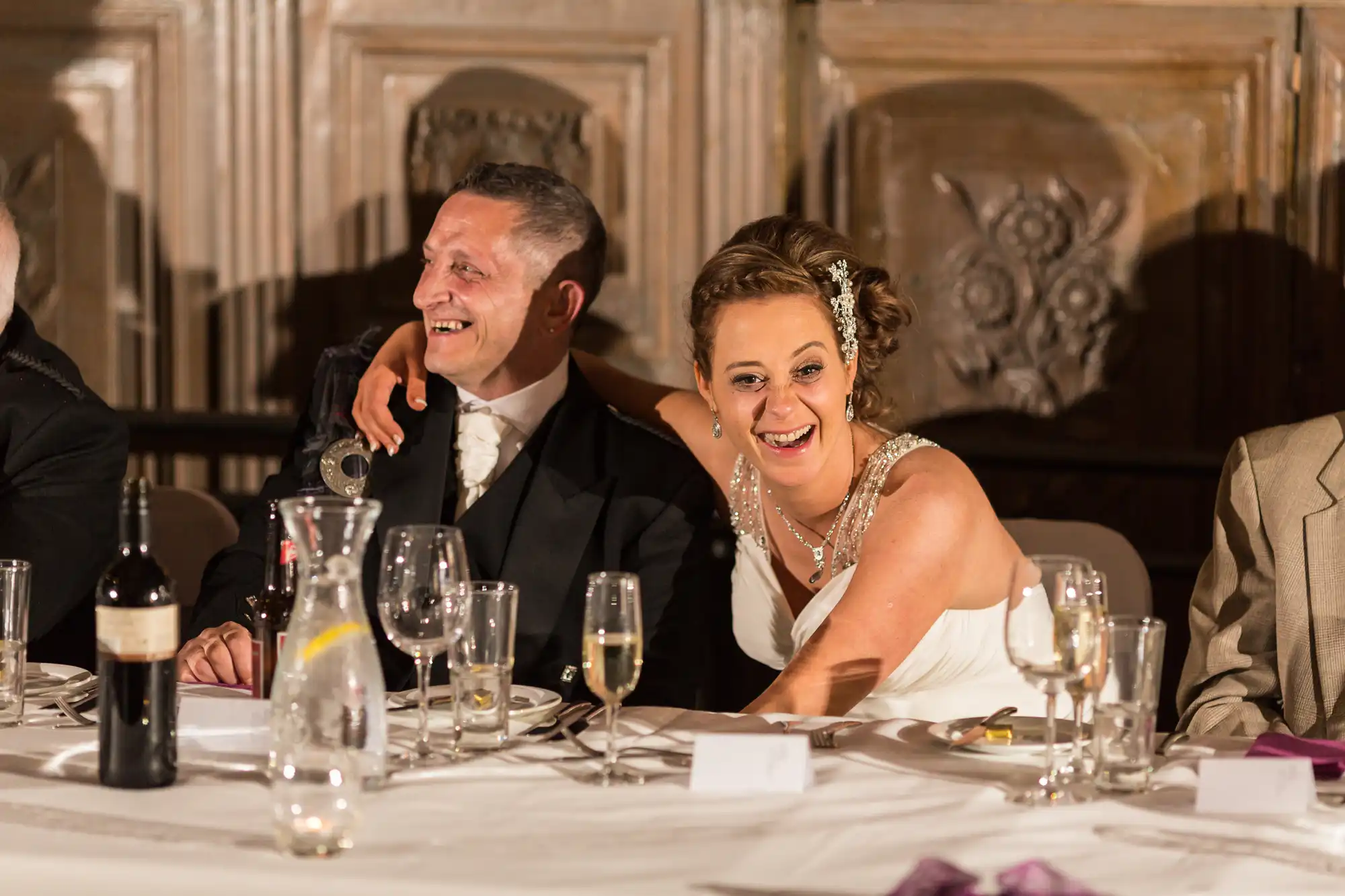 A bride and groom laughing at a wedding reception table, surrounded by wine glasses and decor.