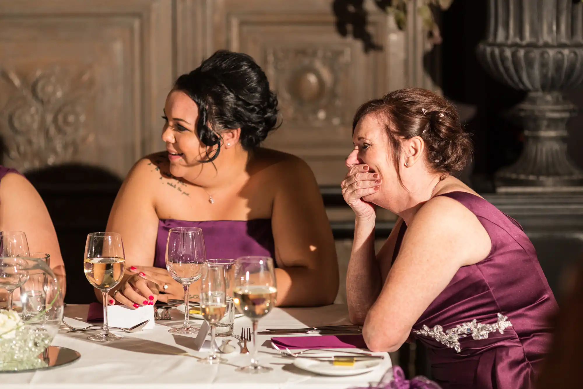 Two women in formal dresses, one in purple, laughing and conversing at a dining table set with wine glasses and white plates in a well-lit elegant room.