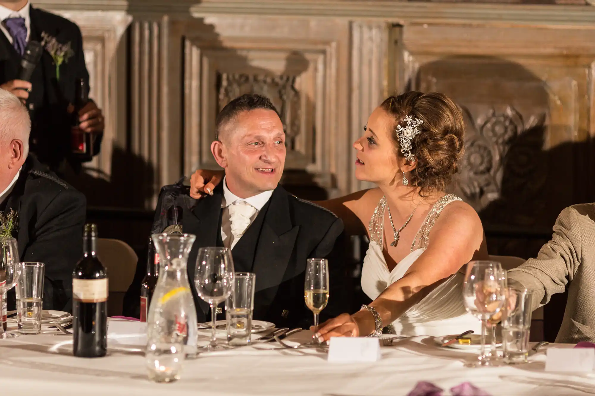 A bride and groom at a wedding reception table, dressed elegantly, looking towards someone off-camera while surrounded by guests and wine bottles.