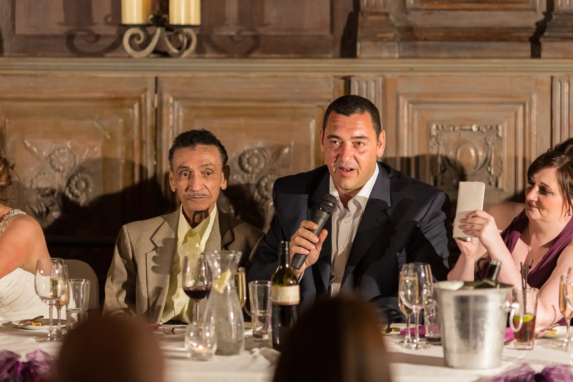 A man in a suit speaks into a microphone at a formal dinner table while others listen, with wine glasses and candles on the table.