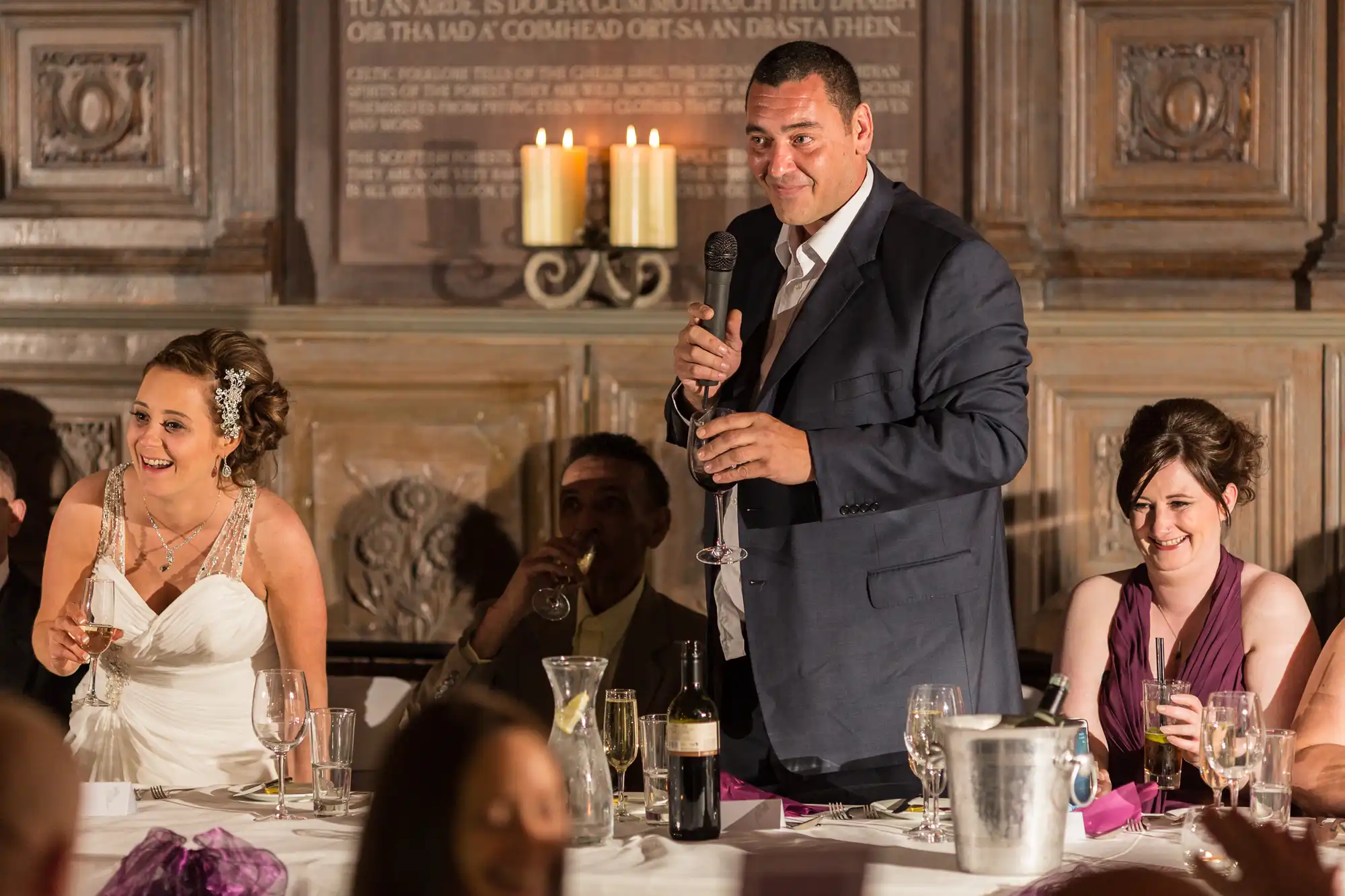 Man giving a speech at a wedding reception, holding a microphone, with a bride and other guests seated and smiling around a dinner table lit by candlelight.