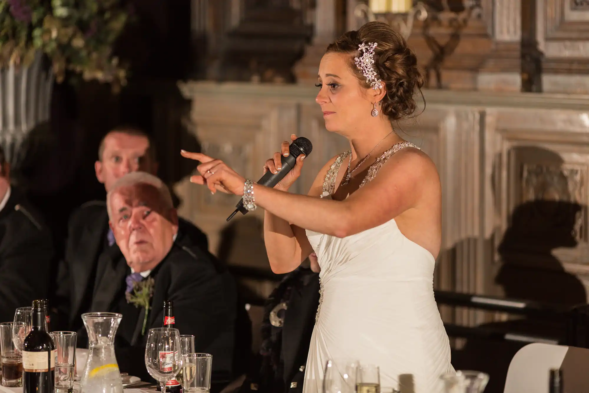 Bride in white dress giving a speech with a microphone, pointing at someone, at a wedding reception indoors.