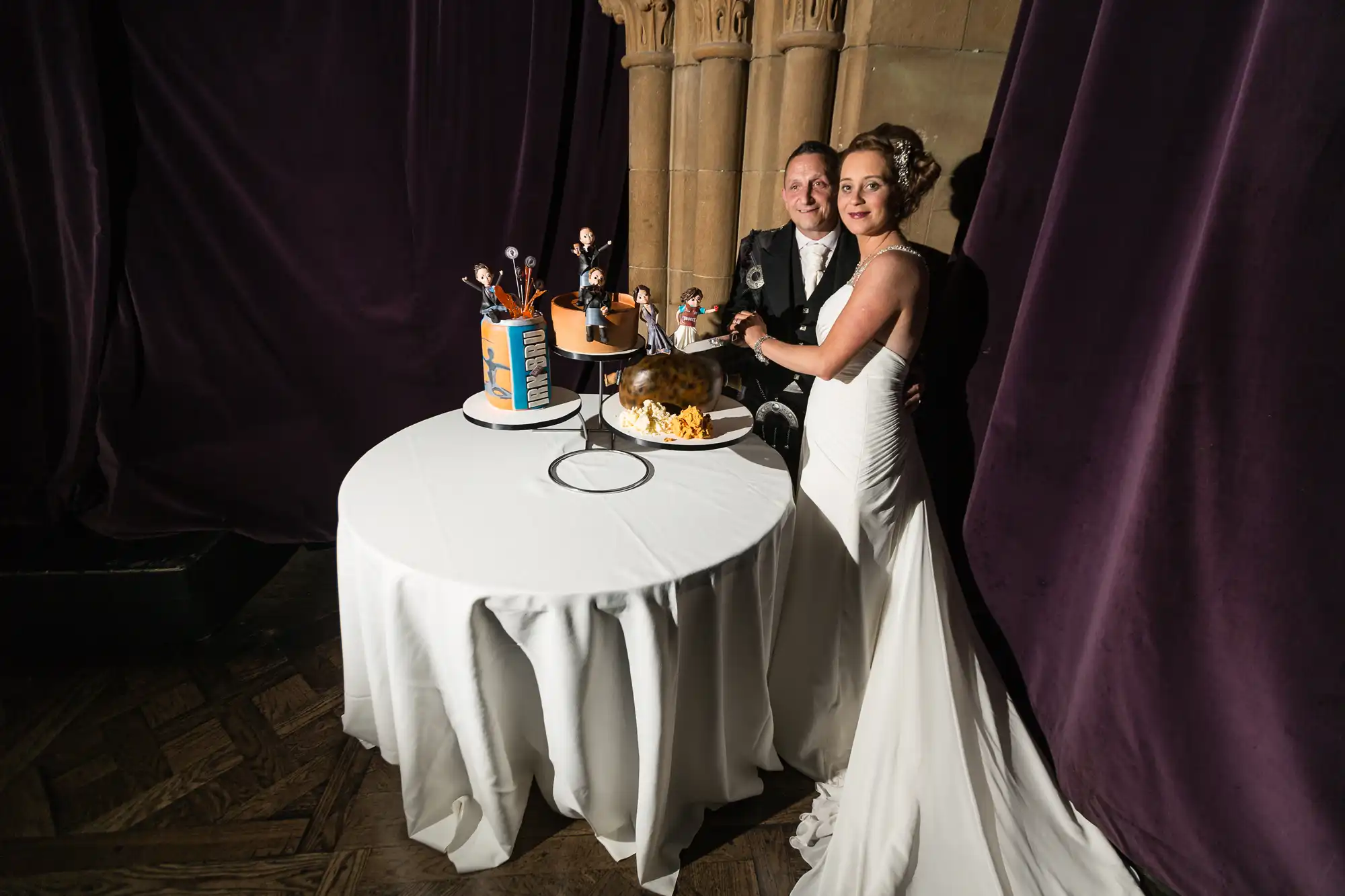 A bride and groom in formal attire cutting their wedding cake at a table, surrounded by curtains in a grand hall.