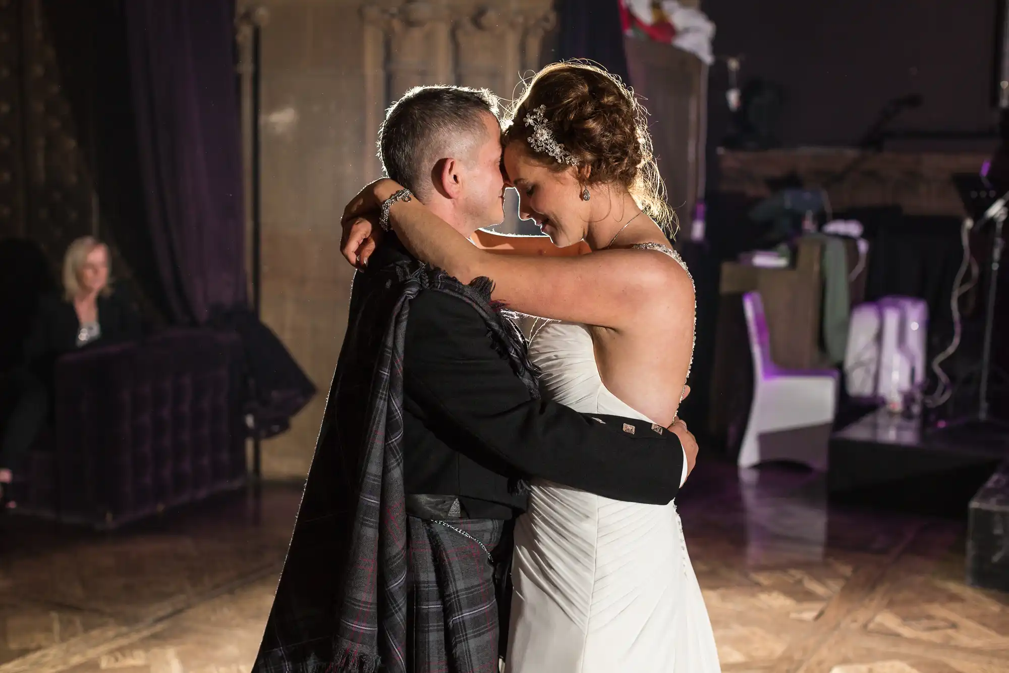 A newlywed couple shares a slow dance, the bride and groom embracing on a dance floor dimly lit with purple lights.