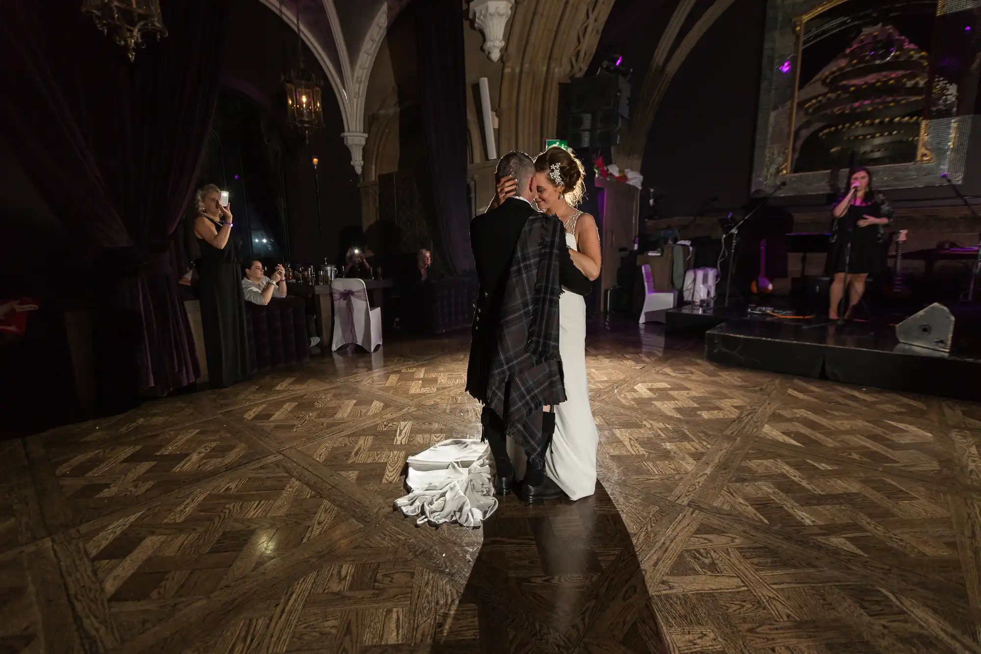 A bride and groom share a dance in a grand ballroom with onlookers and a live band in the background.