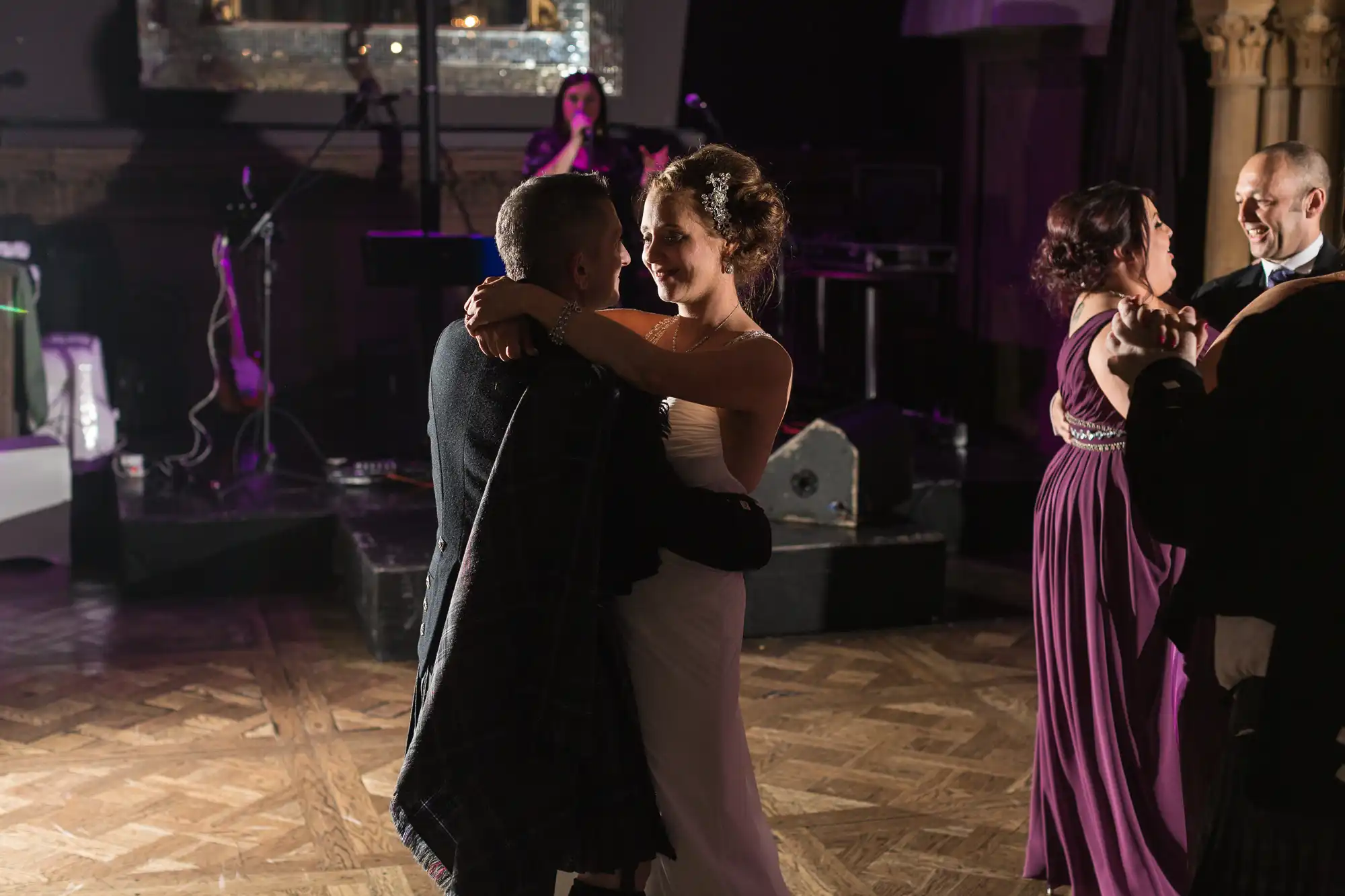 A bride and groom sharing a slow dance at their wedding reception, illuminated by soft lighting, with another dancing couple and a musician in the background.