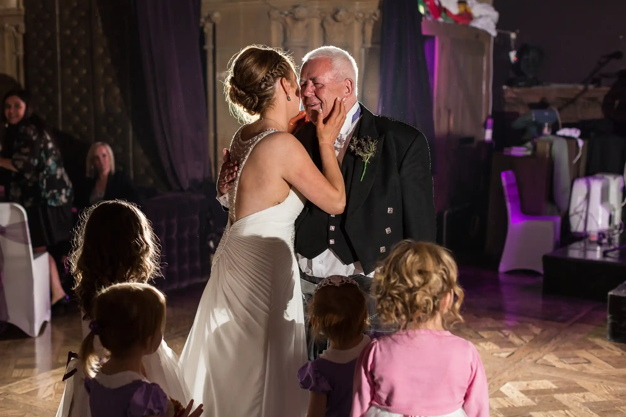 A bride touches the face of an elderly man, both smiling, surrounded by children at a wedding reception.