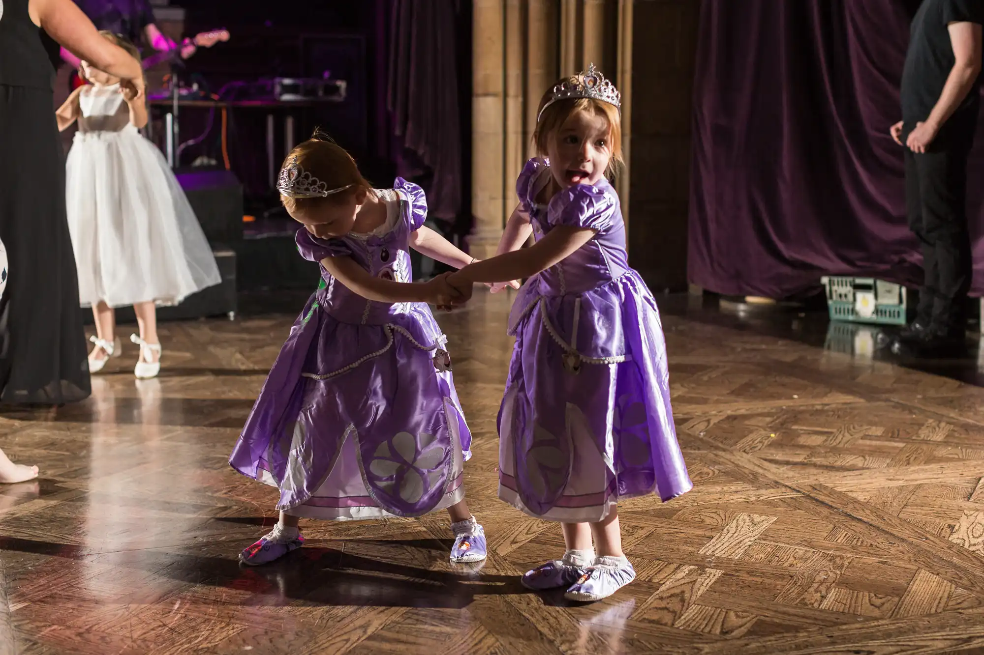 Two young girls dressed as princesses playfully dance on a wooden floor at a formal event, wearing tiaras and purple dresses.