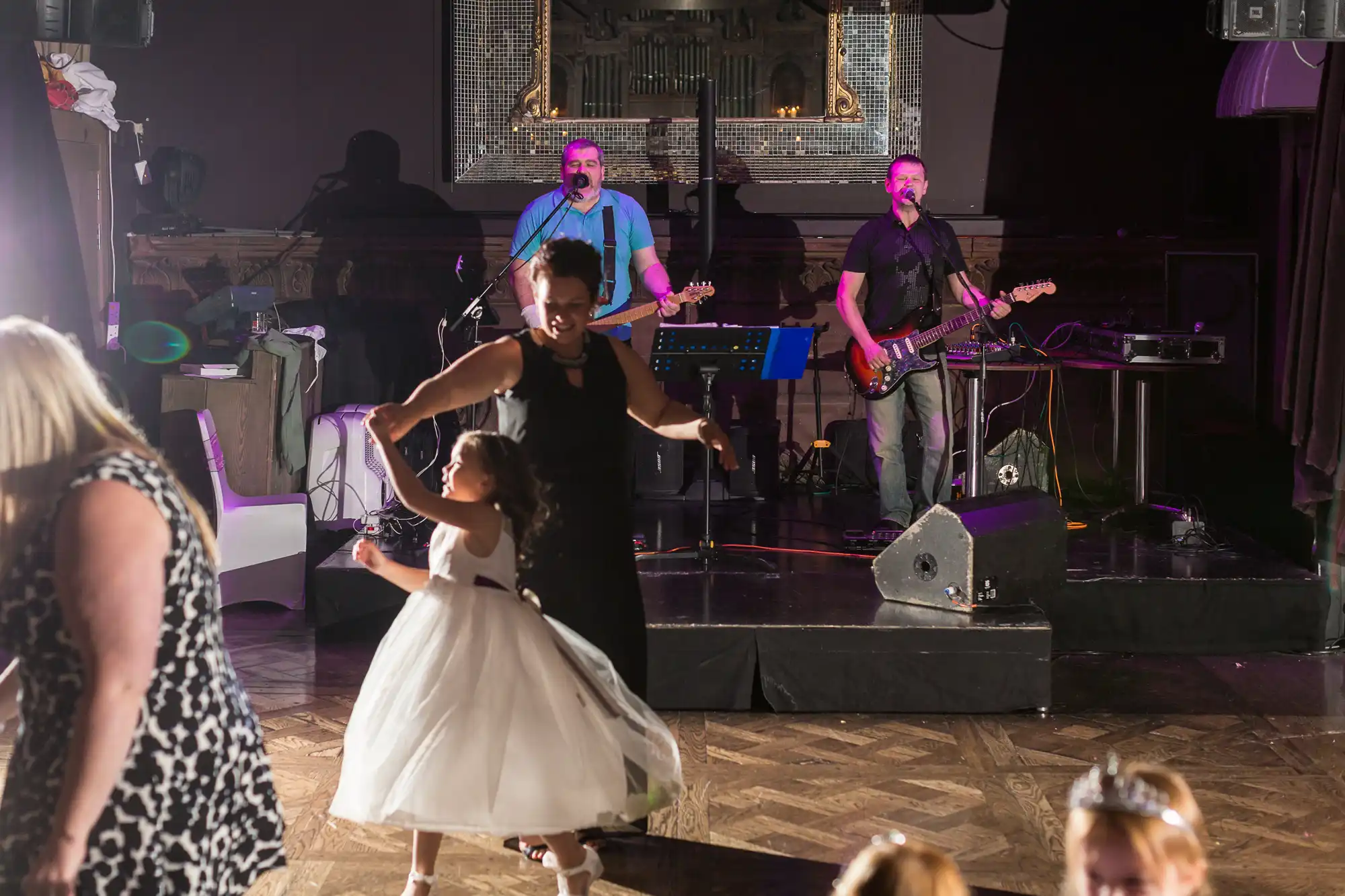 A woman and a young girl dance joyfully at a wedding reception while a band performs on stage in the background.