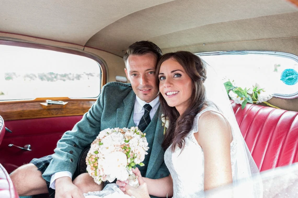 newlyweds smiling as they sit on the back of a classic car