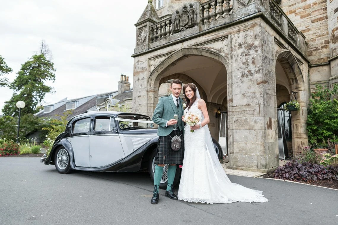 newlyweds holding a glass of champagne standing next to a classic car at the entrance to the Dalmahoy Hotel