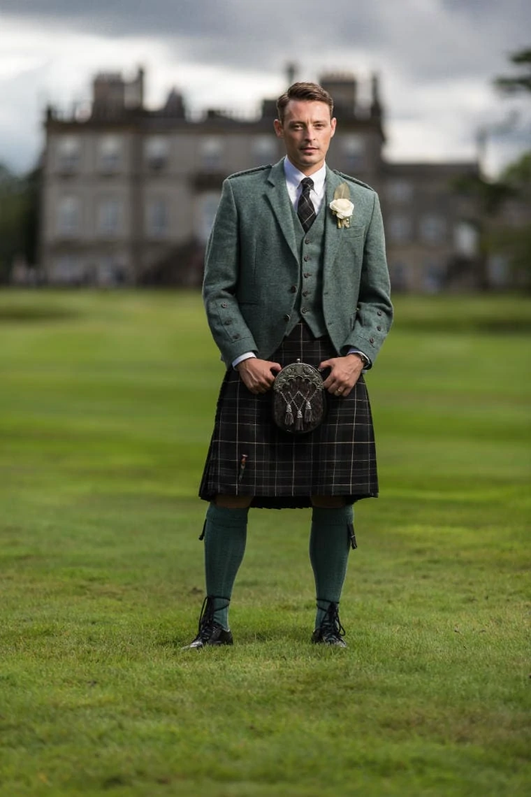groom portrait wearing green tweed kilt outfit and blue tie with the Dalmahoy Hotel in the background