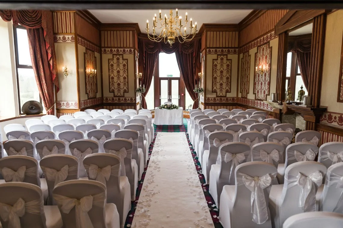Interior of a wedding venue with white chairs arranged in rows, a red carpet aisle, and an ornate chandelier hanging above.