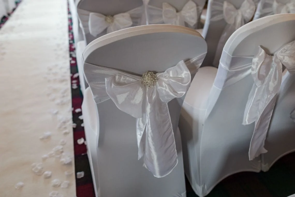 White chairs decorated with satin bows and rhinestone embellishments at a wedding aisle scattered with rose petals.