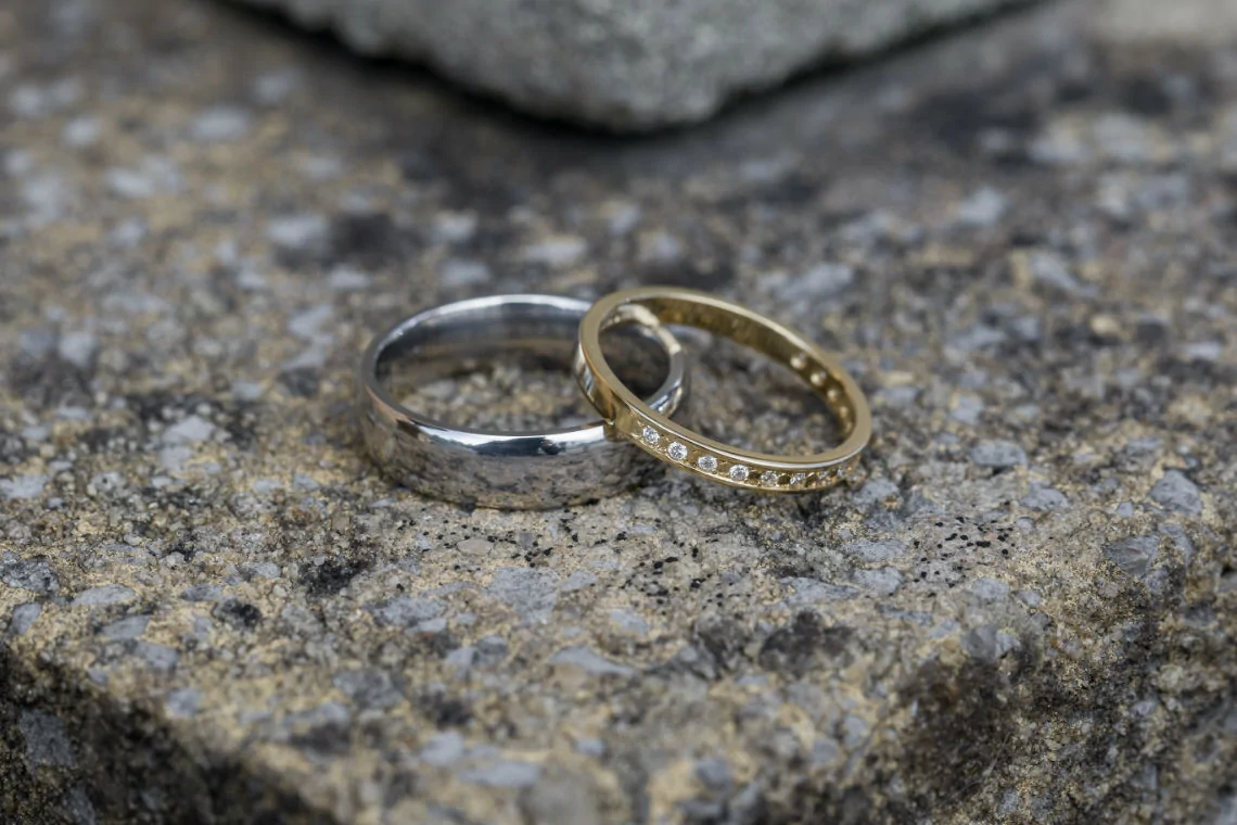 Two wedding rings, one silver and one gold with diamonds, resting on a rough stone surface.