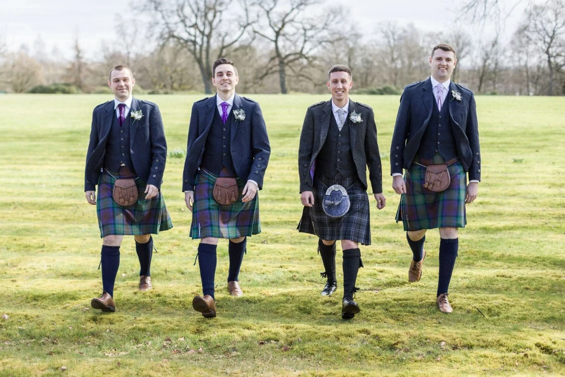 Four men wearing traditional scottish kilts and jackets, smiling and standing side by side in a grassy field.