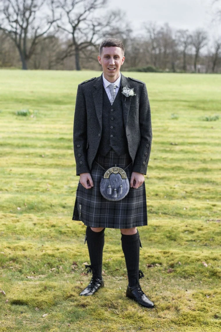 A man in traditional scottish attire, including a kilt and sporran, stands smiling in a grassy field.