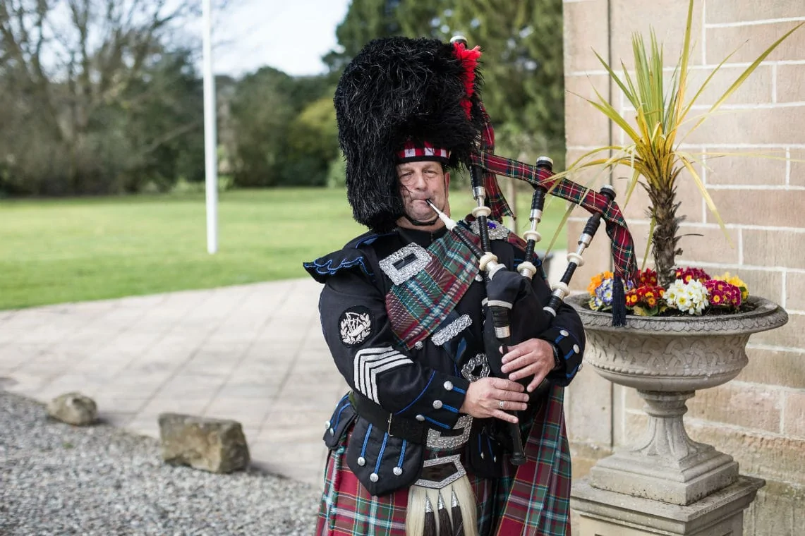 A man in traditional scottish attire, including a kilt and sporran, plays the bagpipes outdoors near a building adorned with potted plants.