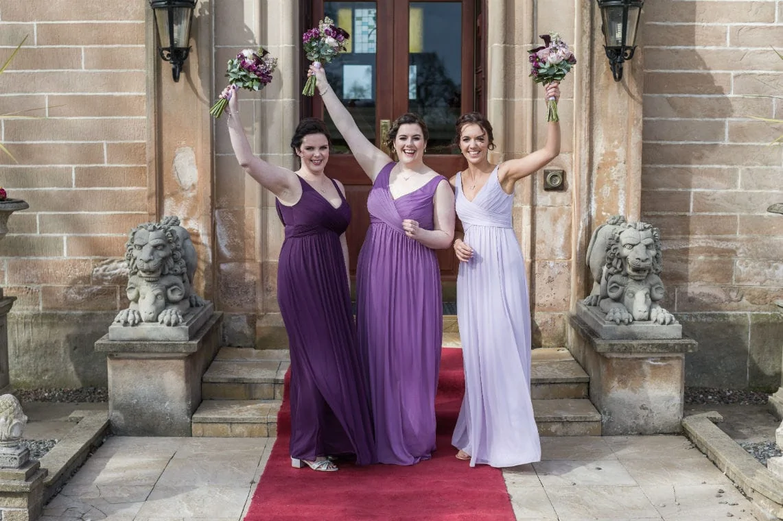 Three bridesmaids in purple dresses, raising bouquets in the air, standing on a red carpet outside a building with stone lion statues.