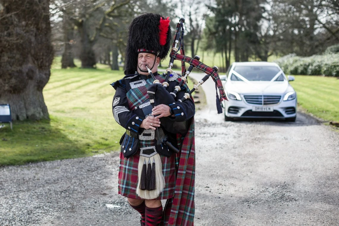A man in traditional scottish attire, including a kilt and sporran, plays bagpipes on a gravel path with a white car in the background.