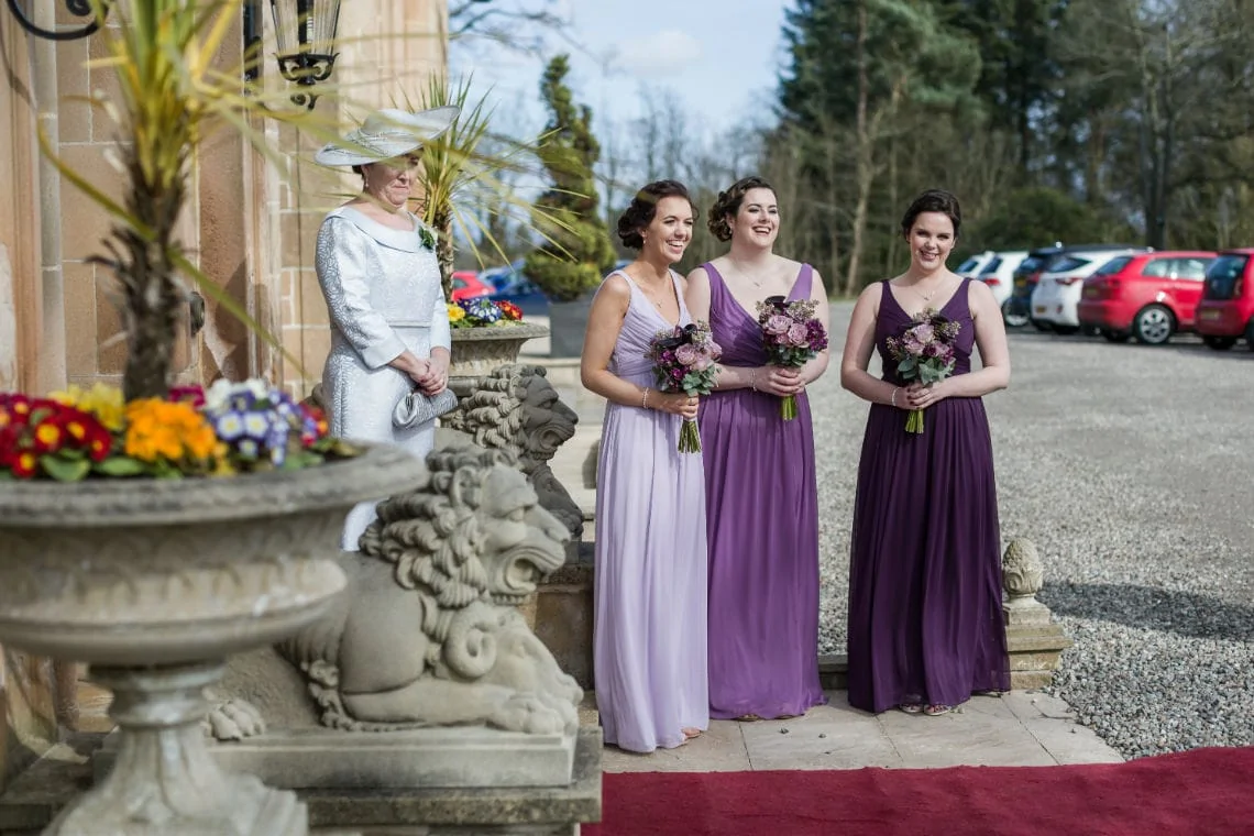 Three bridesmaids in purple dresses holding bouquets stand next to an older woman in a white suit and hat outside a building.