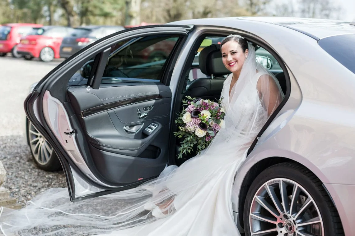 A bride in a white gown and veil, holding a bouquet, smiles from the backseat of a car with the door open.