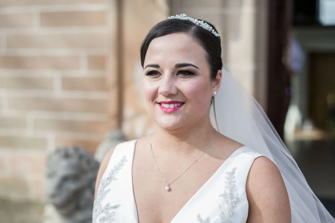 A bride in a white gown with a veil and tiara, smiling outdoors on her wedding day.