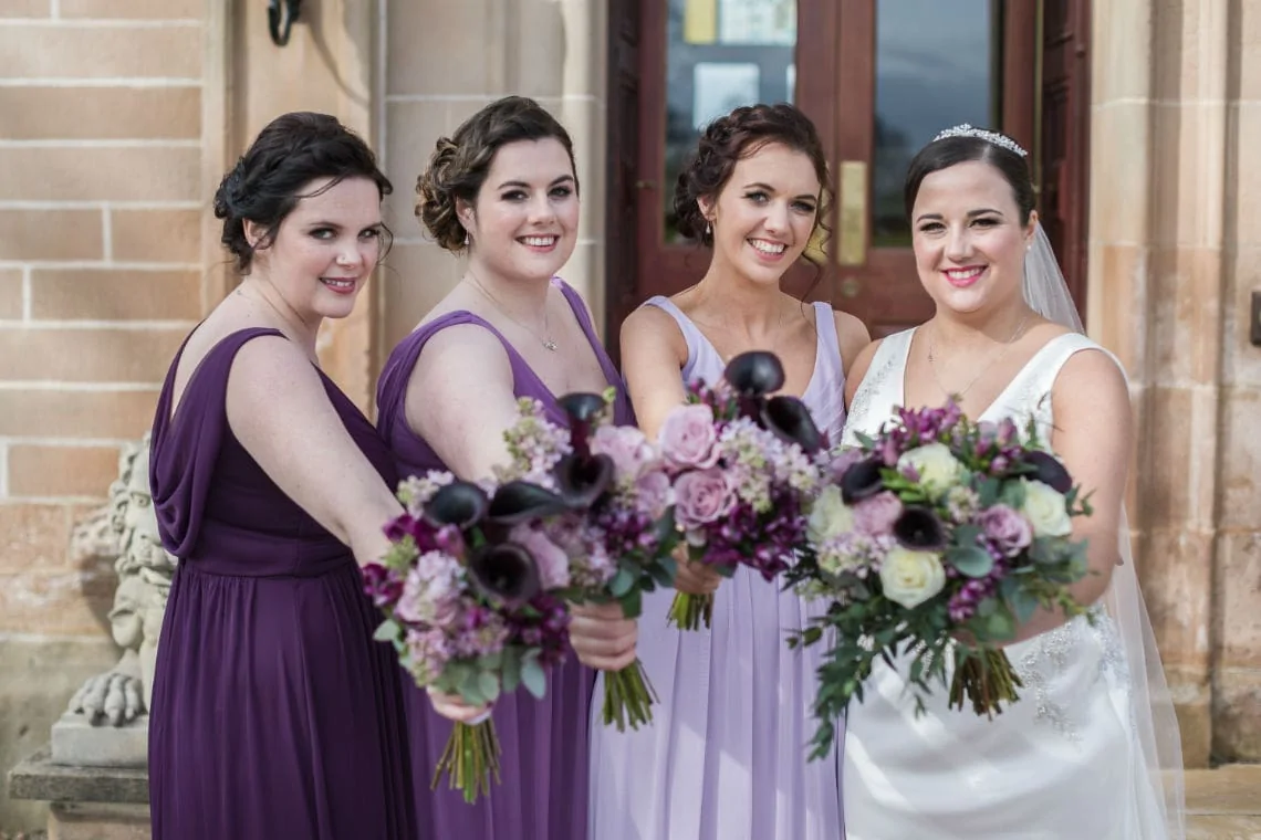 Four women in formal attire, three in purple dresses and one in a white bridal gown, holding bouquets and smiling outside a stone building.
