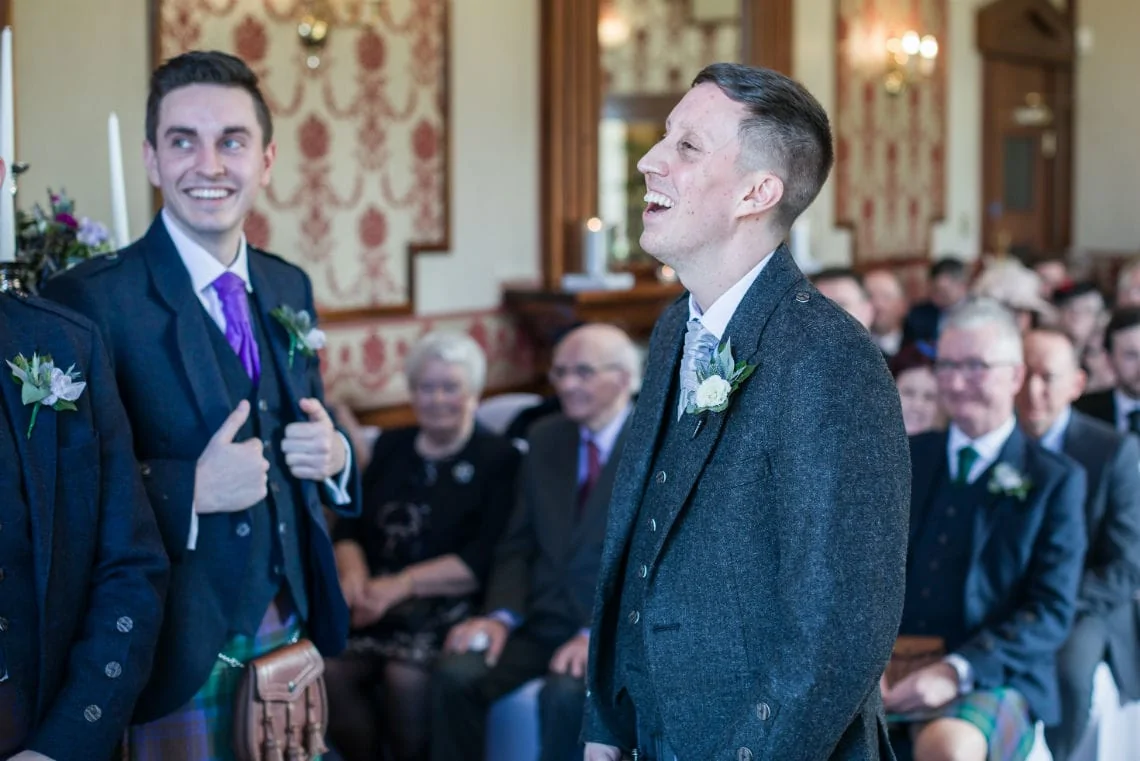 Two men in formal attire, one in a kilt, smiling and gesturing a thumbs-up at a wedding ceremony with guests in the background.