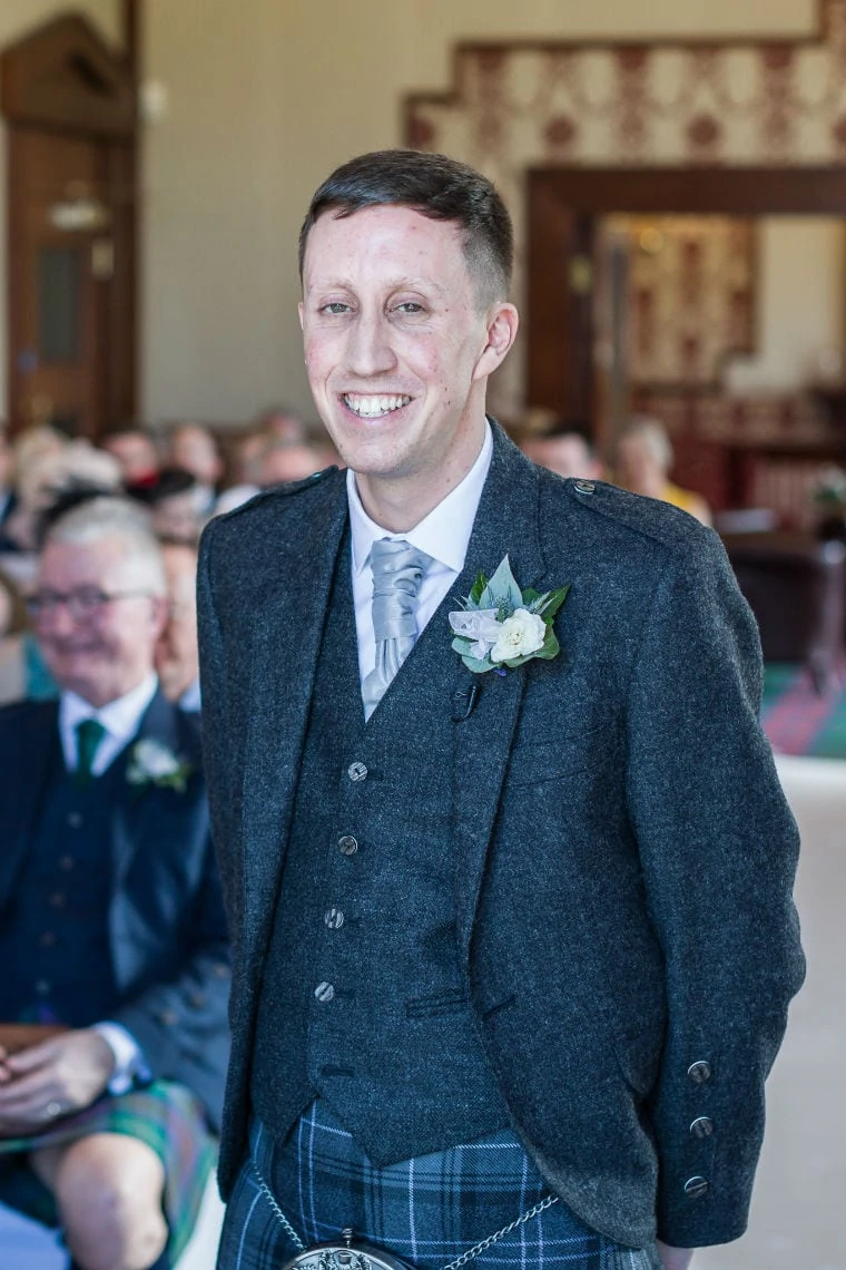 A groom in a gray tweed suit and kilt smiles during a wedding ceremony, with guests visible in the background.