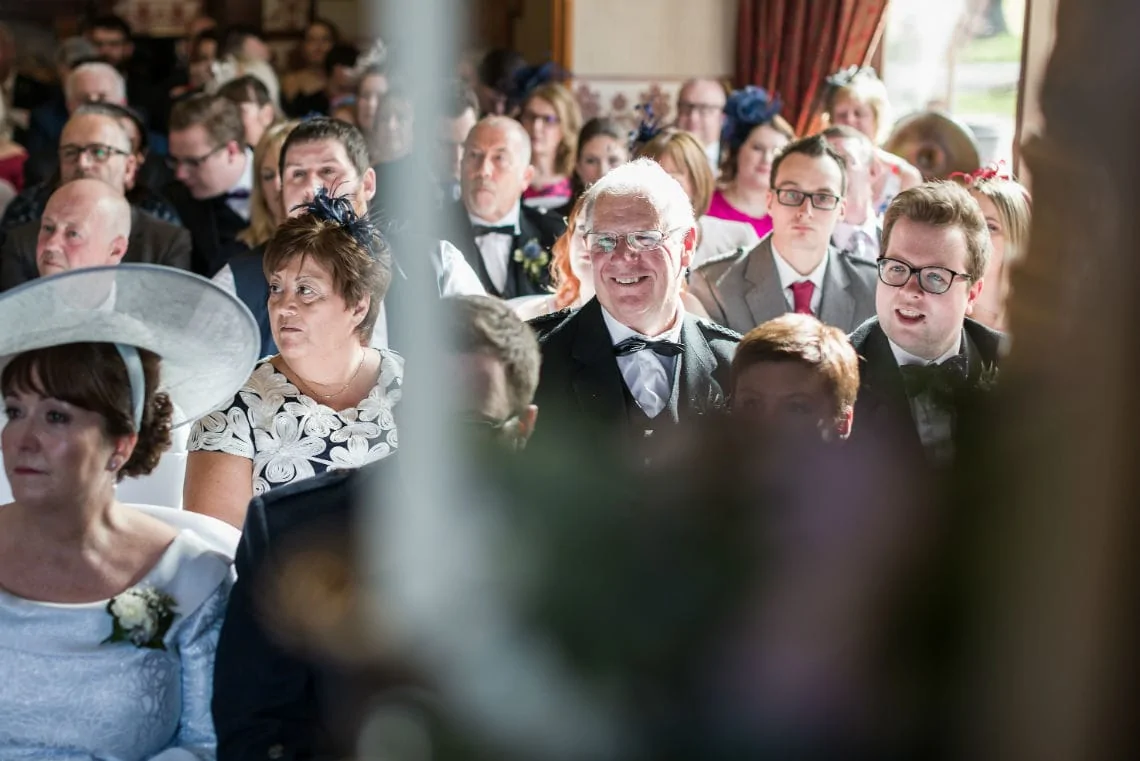 Guests at a wedding ceremony seated indoors, smiling and looking toward the front, with a focus on a middle-aged man wearing glasses.