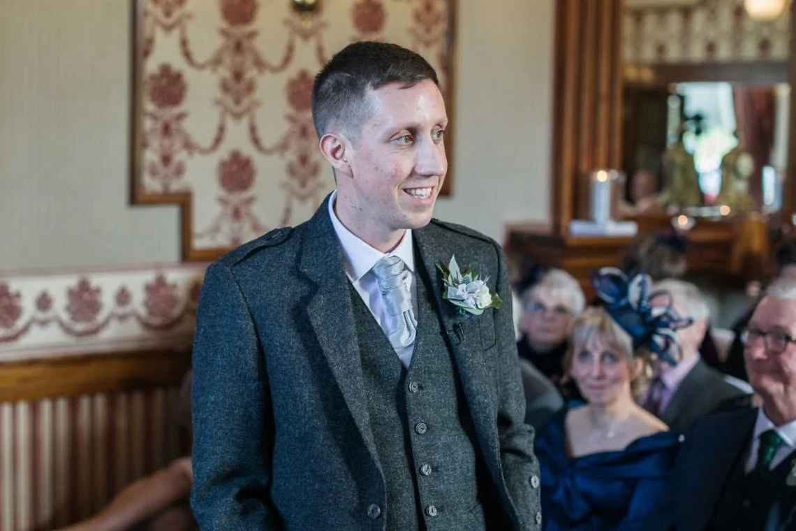 A smiling groom in a grey tweed suit stands in a room with seated guests watching him, adorned in formal attire.