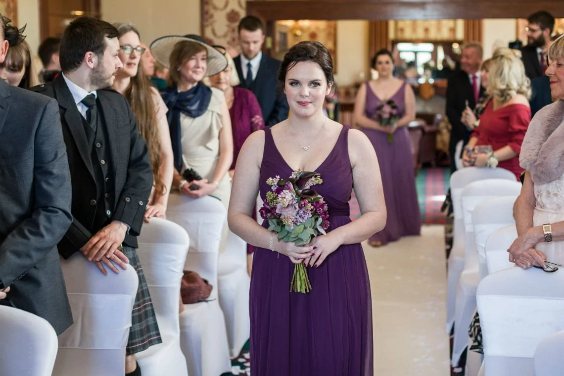 A bridesmaid in a purple dress holding a bouquet stands at a wedding ceremony, surrounded by guests in formal attire.