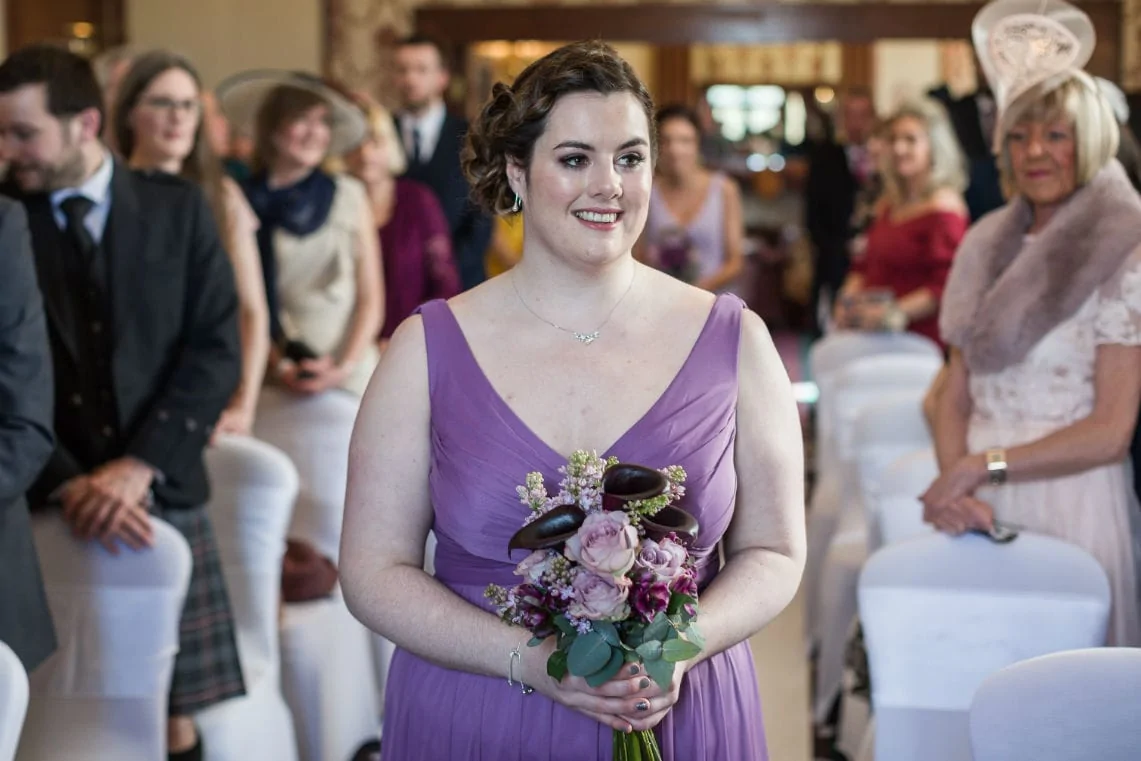 A bridesmaid in a purple dress holding a bouquet stands smiling in a wedding ceremony with guests in the background.