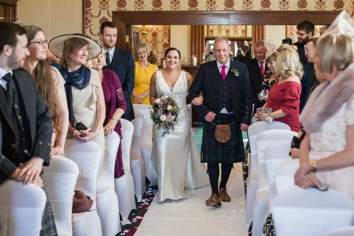 A bride and groom walking down the aisle, guests looking on, groom wearing a kilt in an ornately decorated room.